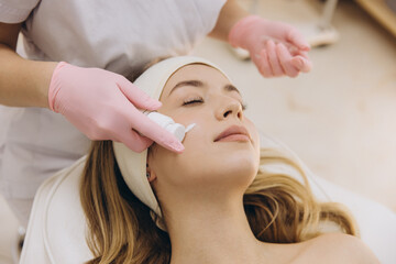 Cosmetologist applying moisturizing cream on face of client in beauty salon for skin rejuvenation...