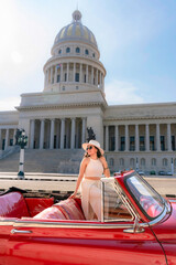 Young Latina woman with sunglasses and a hat smiles while sitting in a vintage red car near El Capitolio in Havana, Cuba.  