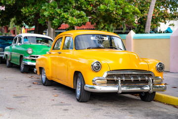 A vibrant yellow classic American car from the mid-20th century parked on a street in Cuba, with another vintage green car behind it.  