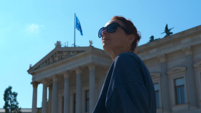 Tourist enjoying sunny day in front of austrian parliament building. Cheerful redhead woman wearing sunglasses relaxing outdoors, gazing upward with austrian parliament building under bright sky