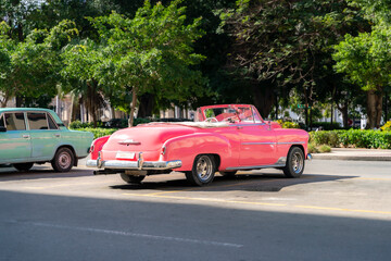 A classic pink convertible from the 1950s parked on a Cuban street, with a green vintage car and...