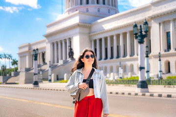Fototapeta premium A stylish woman in sunglasses and a red skirt walks past El Capitolio in Havana, Cuba, on a sunny day with blue skies. 