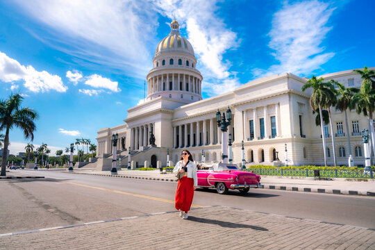 A stylish woman walks across the street near El Capitolio in Havana, Cuba, with a classic pink convertible car and palm trees in the background.  