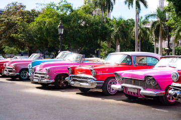 A row of brightly colored classic American cars, including red, pink, and blue convertibles, parked...