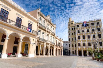 Fototapeta premium A stunning colonial plaza in Old Havana, Cuba, features elegant historic buildings with arched corridors, intricate balconies, and Cuban flags under a bright blue sky. 
