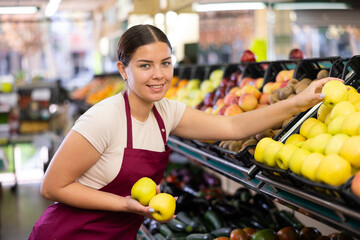 Young girl seller has chosen ripe fruits and holds three shiny sweet apples in hands.Kiwis and apples in boxes in background