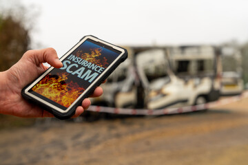 Insurance scam artist holding smartphone with burnt camper van in background
