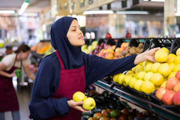 Young woman seller in hijb and apron sells apples in grocery store