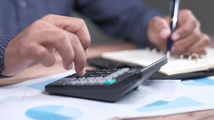 Black man office worker in a suit counts on calculator and records the results with a pen in paper tablet while sitting at desk with laptop