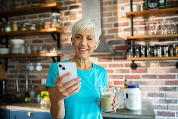 Senior woman drinking protein shake and using smartphone in kitchen