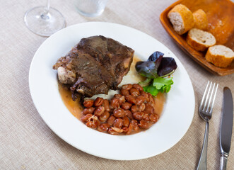 Delicious baked pork cheek served for dinner on white plate with side of spicy stewed pinto beans, garnished with fresh greens. Traditional Spanish comfort dish