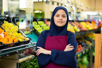 Young woman seller in hijab posing near shelves with vegetables and fruits in grocery store