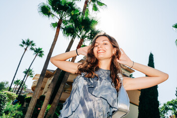 A young woman in a flowing gray dress smiles brightly, walking among palm trees with a modern building in the background