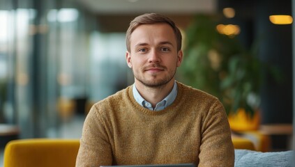 Fototapeta premium Young man with light brown hair and neutral sweater sitting at the table with laptop in modern indoor workspace smiling and looking directly at the camera