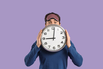 Handsome young man with sleep mask and clock on lilac background
