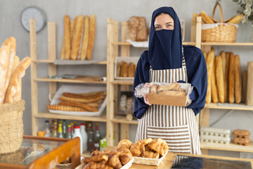Hospitable young saleswoman of European descent dressed in blue niqab and striped apron presenting...