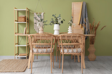 Wicker armchairs and dining table with eucalyptus branches in vases near green wall in room