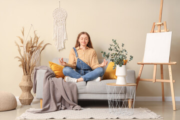Young woman meditating while sitting on sofa near easel with painting in room