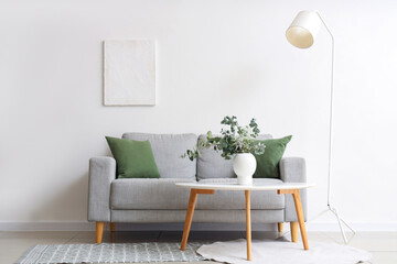 Cozy sofa, coffee table with eucalyptus branches in vase and lamp near white wall in room