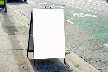 A blank white A-frame sandwich board sign stands on a concrete sidewalk next to a bicycle lane,mockup for street advertising or business messaging.Empty display sign with customizable copy space.