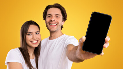 Handsome guy holding and showing smart phone with black empty screen in hand close up to camera, mock up macro, young lady embracing man, family recommending app on mobile phone, selective focus