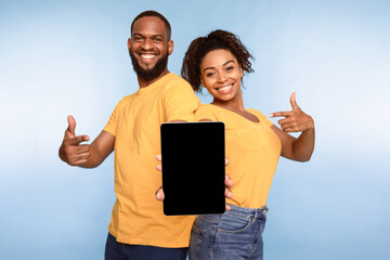 Cheerful African American couple holding tablet pc together, pointing at empty screen with space...