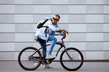 Obraz premium Portrait of handsome black guy with backpack and sunglasses riding bicycle near brick wall outside. Attractive African American hipster traveling in city, going for ride on bike