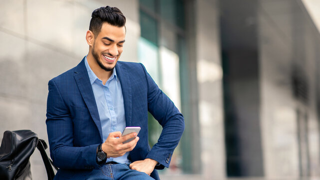 Wealthy middle-eastern entrepreneur using cellphone, sitting on the street by office building with backpack and laptop, resting after business event, checking emails, using business app, copy space