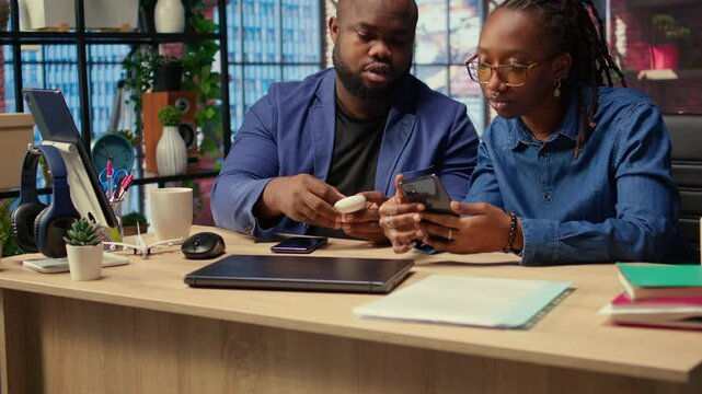Young black couple sharing ear buds to listen to webinar on smartphone, working together on their freelancing tasks at home. Man and woman discussing about a work project. Camera A.