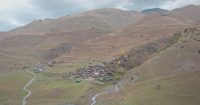 The drone approaches a historic village nestled among steep slopes in the Georgian highlands. The traditional settlement of wooden and stone houses is surrounded by narrow paths and a small stream.