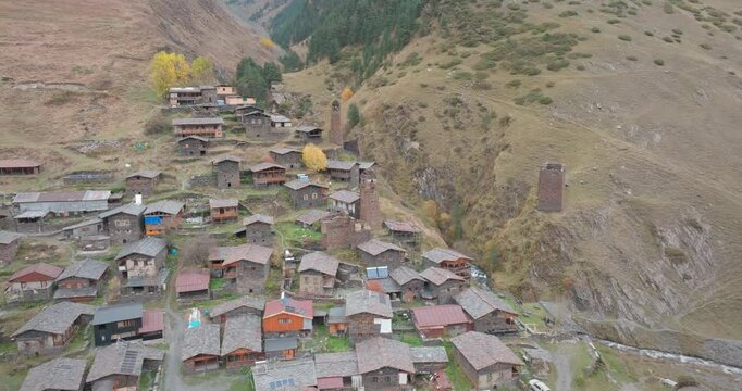 The drone circles the village, surrounded by historic stone houses and defensive towers in the mountainous area. The steep valley and autumn colors add depth to the landscape.