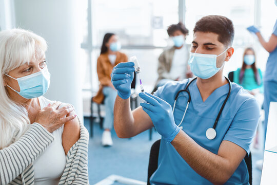 Young doctor in medical face mask and uniform drawing medication out of vial preparing syringe before injecting mature woman with flu or flu vaccine in arm during mass vaccination campaign
