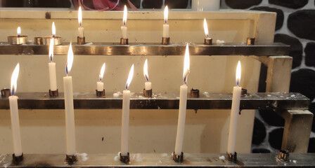 lit candles on metal stands are arranged in two rows, with small candles in the top row and large candles in the bottom row, in low light condition, background includes a stone wall pattern, peaceful