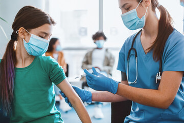 Nurse or doctor in medical face mask giving flu injection to little patient during mass immunization campaign at health center. Schoolgirl getting flu vaccine shot at clinic or hospital