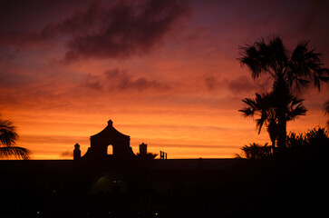 A striking silhouette of a building with palm trees against a vivid orange and red sunset sky, creating a dramatic scene.