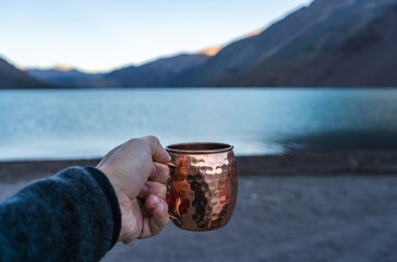 Hand holding a copper mug by a mountain lake at sunrise. Relaxing outdoor moment, perfect for travel, camping, adventure and lifestyle concepts.