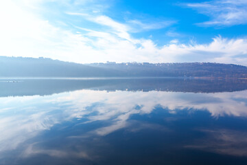 Serene lake with a clear reflection of sky and distant hills on its calm surface. Sky is partly cloudy with wispy clouds, and the horizon features a line of buildings or structures atop the hills