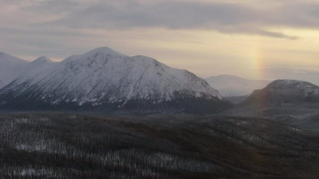 Experience a breathtaking golden hour aerial shot over snow-covered Ibex Valley in Yukon. Mountains and dense forests emerge from a sub-zero winter landscape, embodying a true northern wonder.