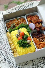A beautifully arranged Indonesian tumpeng box or nasi dus with yellow rice, fried chicken, noodles, and side dishes, garnished with quail eggs, tomatoes, and fresh herbs on a lace tablecloth.