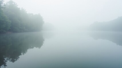 Fototapeta premium A calm lake with fog floating over its surface, reflecting the surrounding trees and creating a meditative atmosphere.