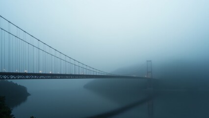 Obraz premium A dramatic suspension bridge enveloped in fog, with a quiet river below, creating a moody and atmospheric landscape.