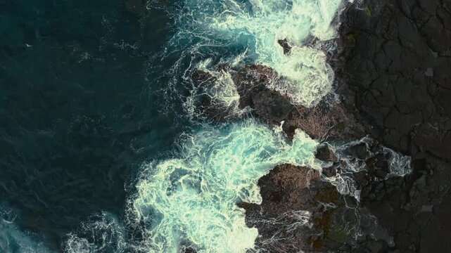 Powerful ocean waves collide with jagged rocks, creating dramatic white foam patterns in this aerial view of the coastline.