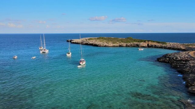 Breathtaking view of several sailing boats anchored in the vibrant turquoise waters of Cala Varques. The sun shines brightly over the stunning coastline of Mallorca, creating a serene atmosphere.