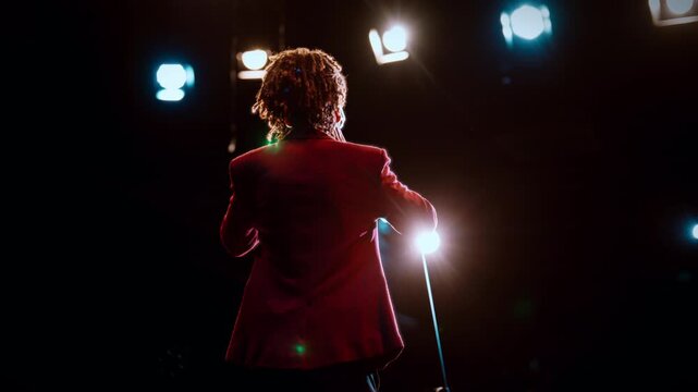 Successful black man speaking on stage, back view, stand-up comedy show. Rearview silhouette in bright soffits of young adult man with dreadlocks speaking for audience, presenting marketing strategy