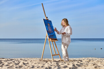 Young woman artist stands near ocean, painting on easel. Serene beach environment complements her...