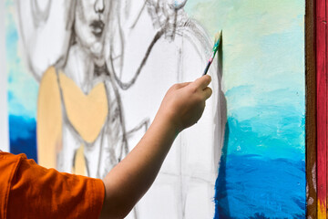 Young caucasian male child in orange shirt painting mural outdoors. Bright sunlight enhances his focused expression. Blue and yellow dominate background © TRAVELARIUM