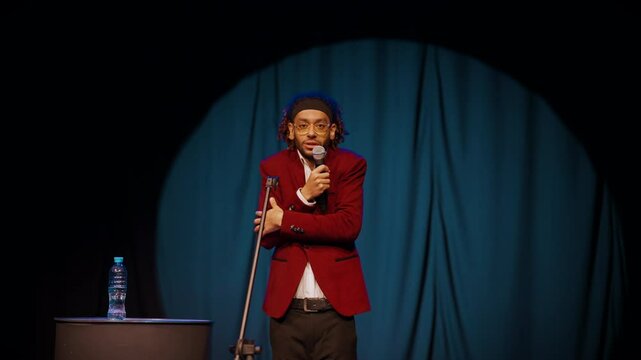Handsome Arabic man speaking stand-up monologue on stage, medium portrait. African american male person performing in show, black guy with professional microphone, comedy performance, slow motion