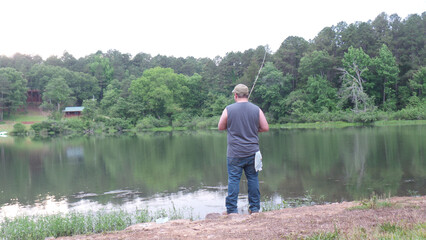 Man fishing by a lake with forested hills and cabins in the background
