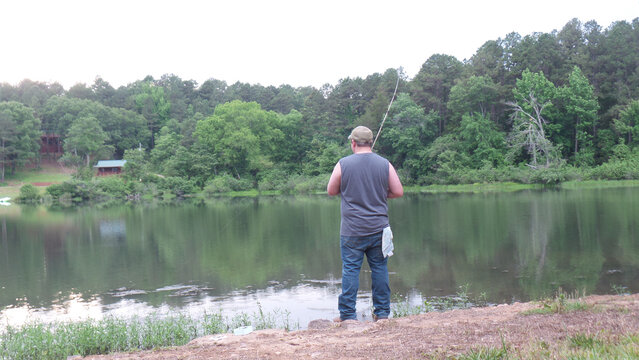 Man fishing by a lake with forested hills and cabins in the background
