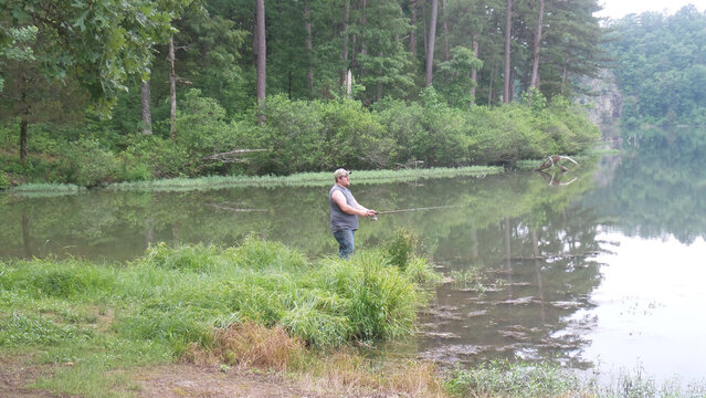 Man fishing by a calm lake surrounded by lush greenery and trees